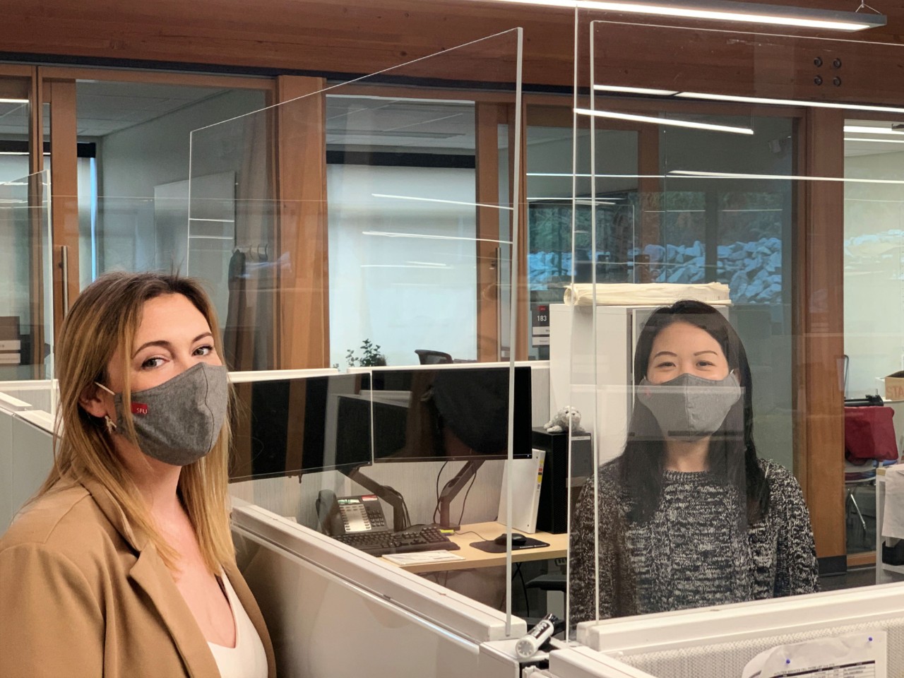 Krystal Ness and Novia Chow look at the camera. Ness is standing and Chow is sitting at her computer desk; both are wearing masks. There is a tall plexiglass barrier between them.
