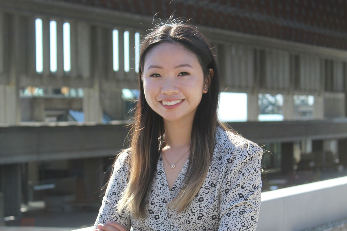 Joelle Mariano smiles at the camera, standing on one of the balconies in SFU's Academic Quadrangle. She is wearing a black and white patterned top.