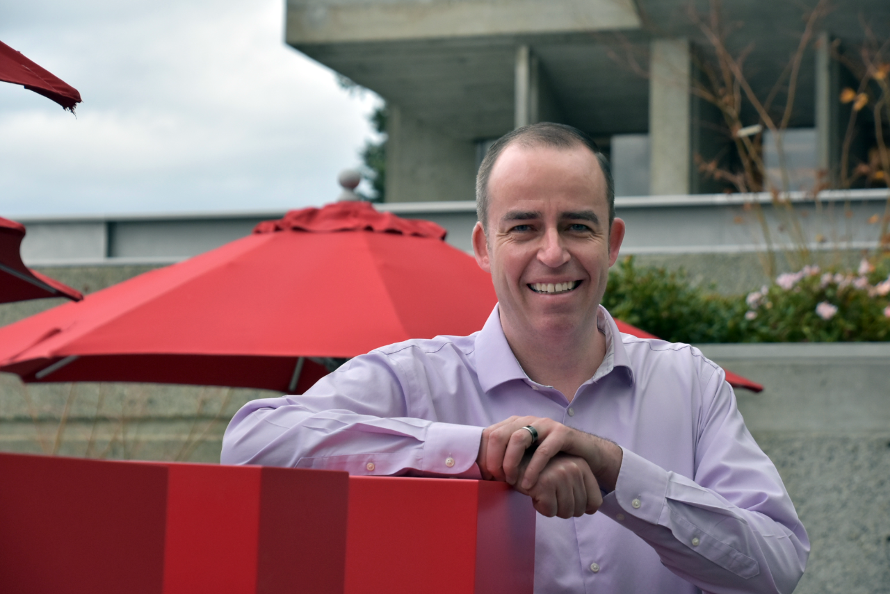 Tom Nault leans on a red post, smiling at the camera. He is wearing a purple shirt, and SFU's Burnaby campus—including a red umbrella and the AQ—are visible in the background.