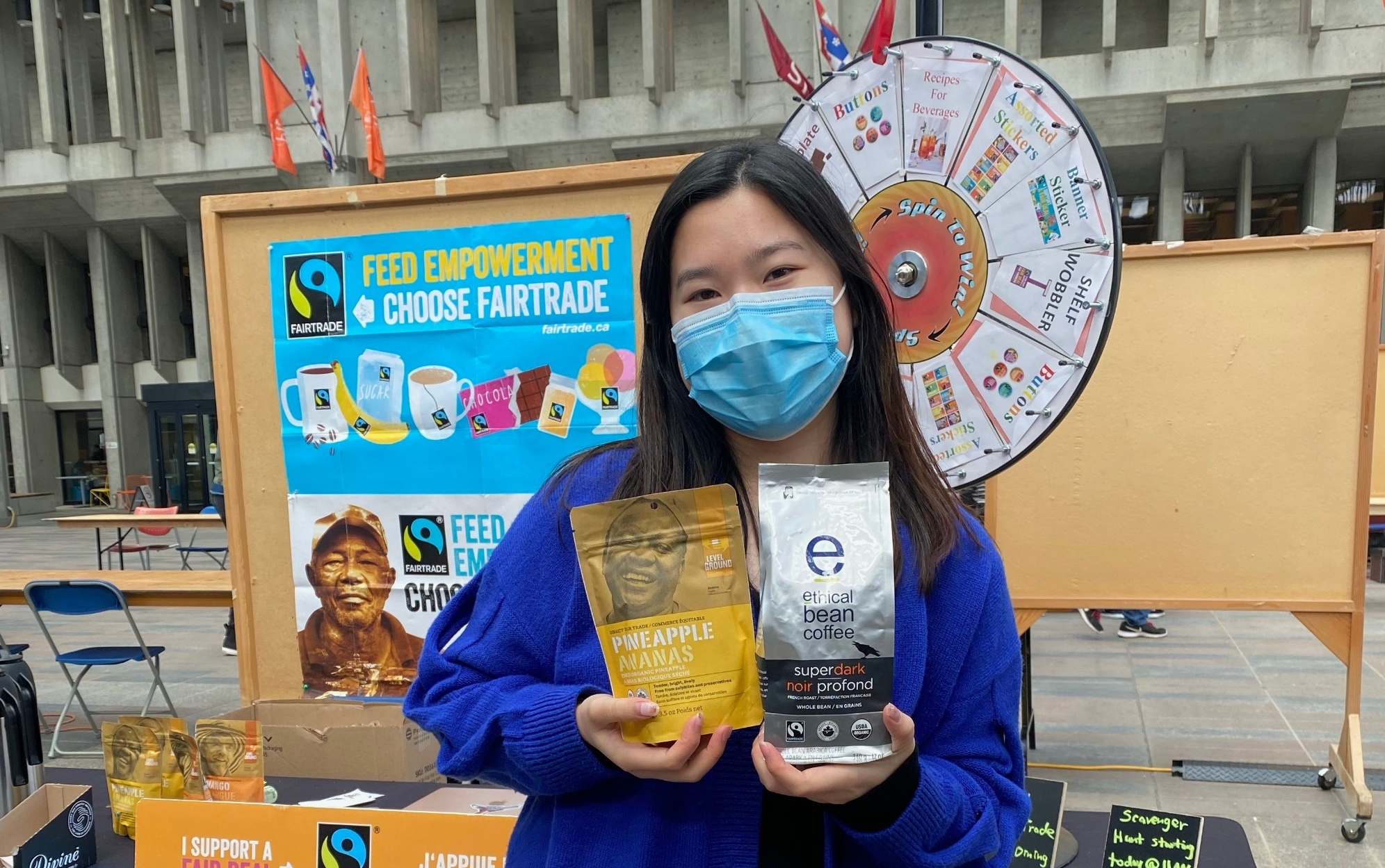 A person with long dark brown hair wearing a medical mask and holding two fair trade products in front of a table and poster board with fair trade awareness materials on them in Convocation Mall.