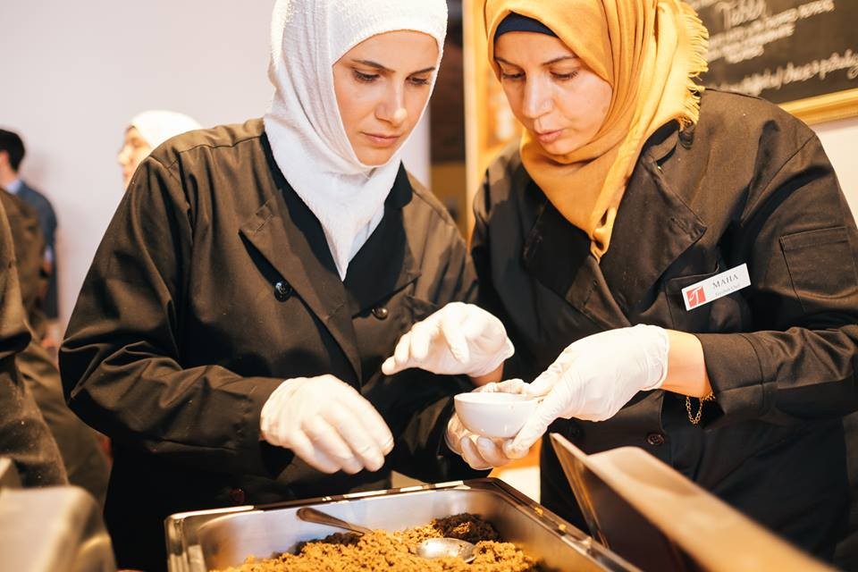 two people wearing hijab adjusting a serving platter of Syrian food