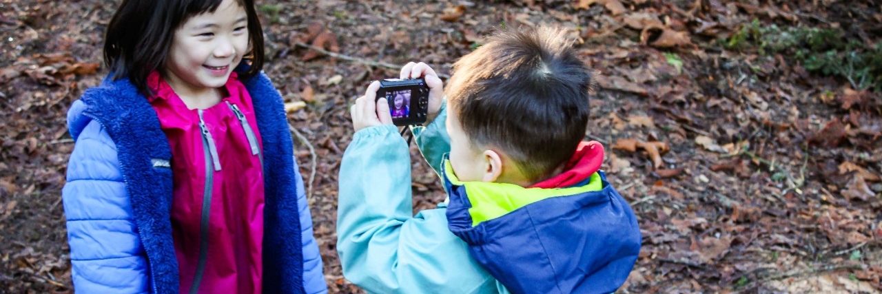 Image is of two children hanging out outside. one is taking a picture of the other who is smiling. Both children are wearing their raincoats and having fun