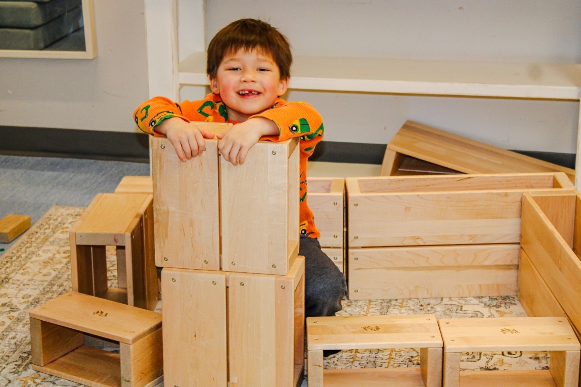 a child in the 3-5 program is facing the camera smiling as they play with blocks and other wooden objects
