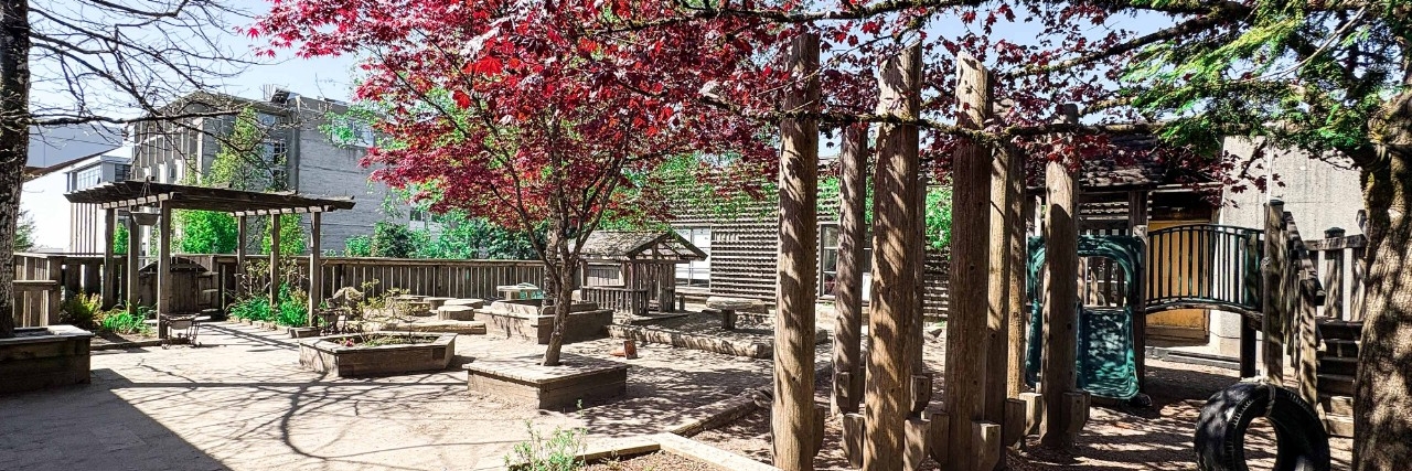 Image is of a typical play areas of SFU Childcare on the Burnaby Mountain west side of campus. There is a red tree in the middle, as well as a picture of a treehouse with a slide and play area
