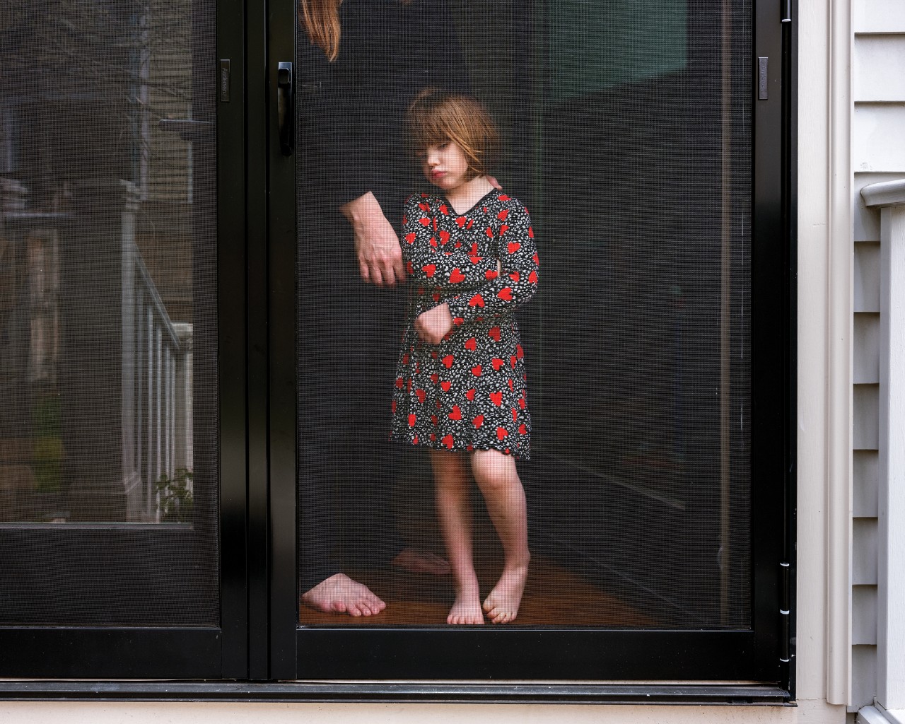 Figure 6. “Ellie and Megan, Cambridge, Massachusetts, 2020.” Ellie, a small child clothed in a floral dress, stands in front of her partially shown mother, Megan, as both look out their front door window. 