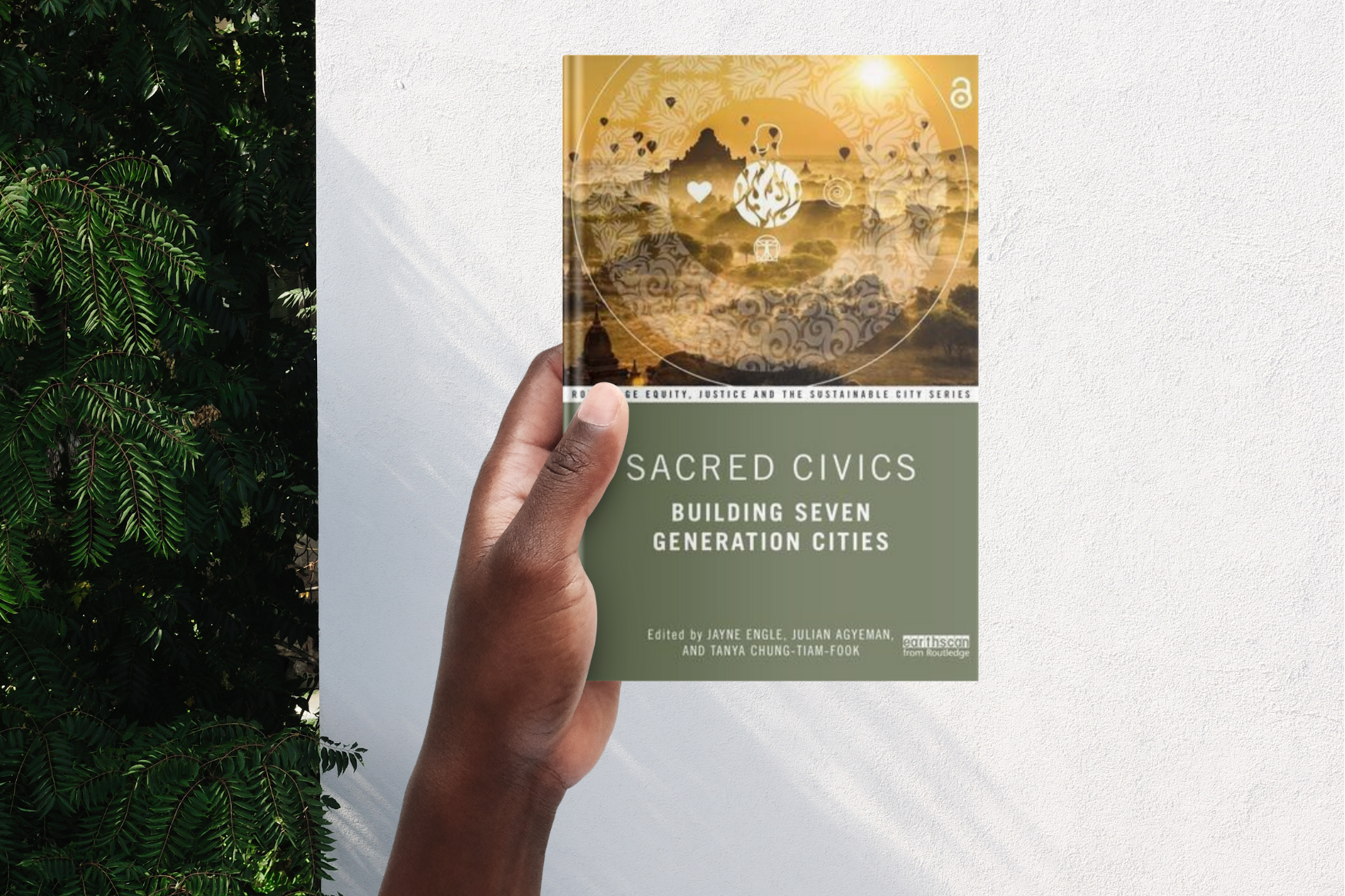 Hand holding the "Sacred Civics" book in front of a white wall with a green plant in the background