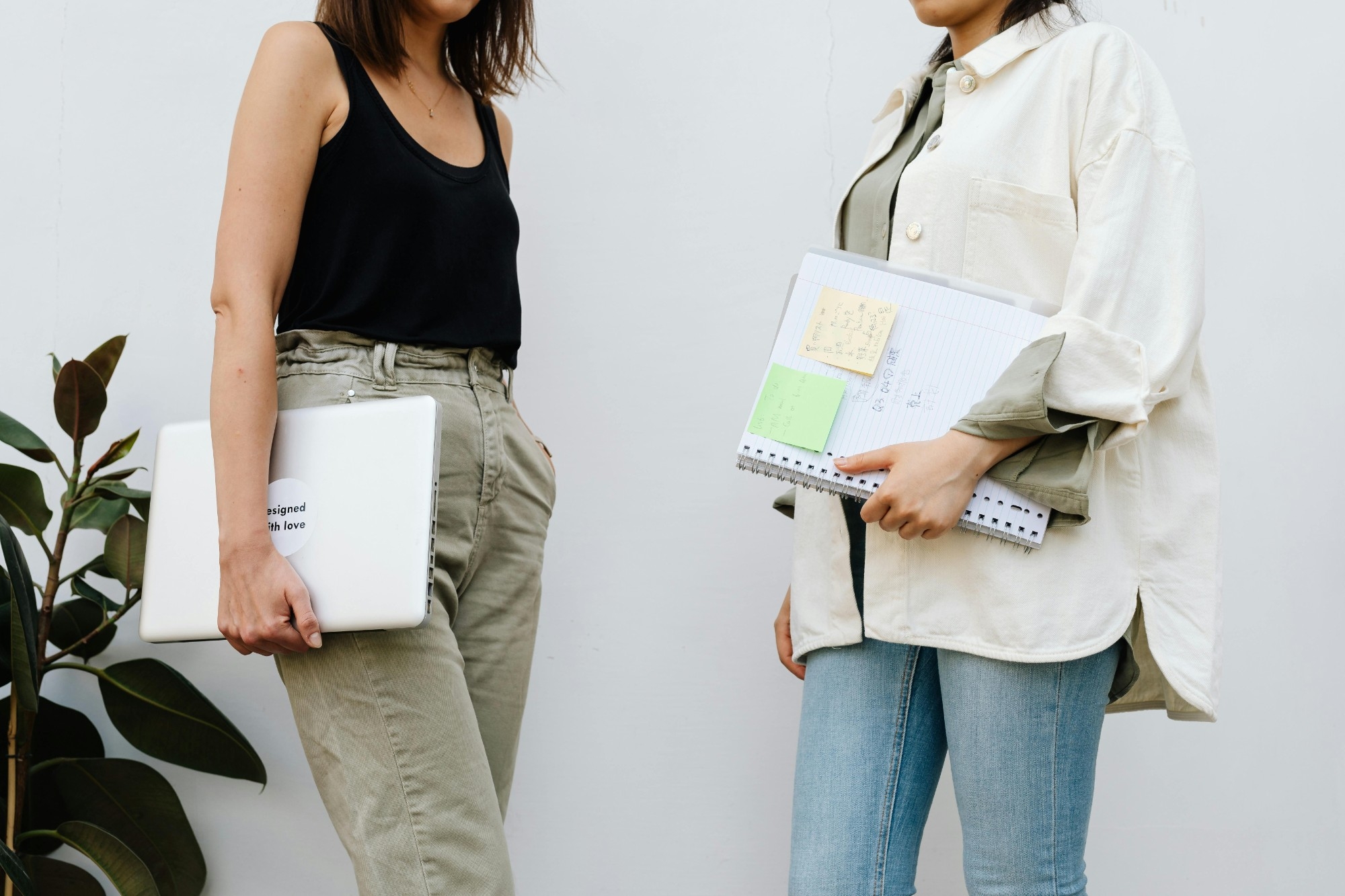 Two dialogue practitioners standing in an office, one holds a laptop and the other holds a notebook with sticky notes on it, the image is cropped below their faces so only torsos are seen