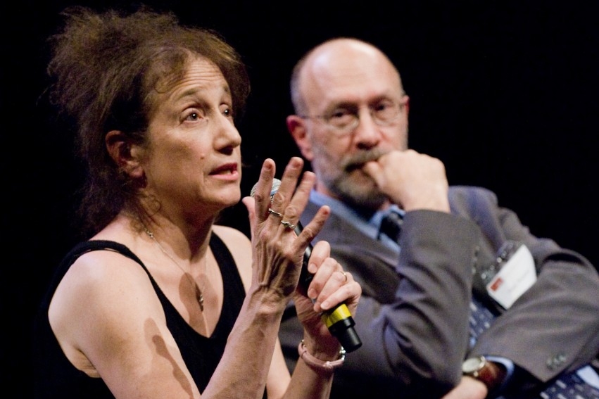 Liz Lerman speaking with a microphone, Mark Winston listening and sitting beside her