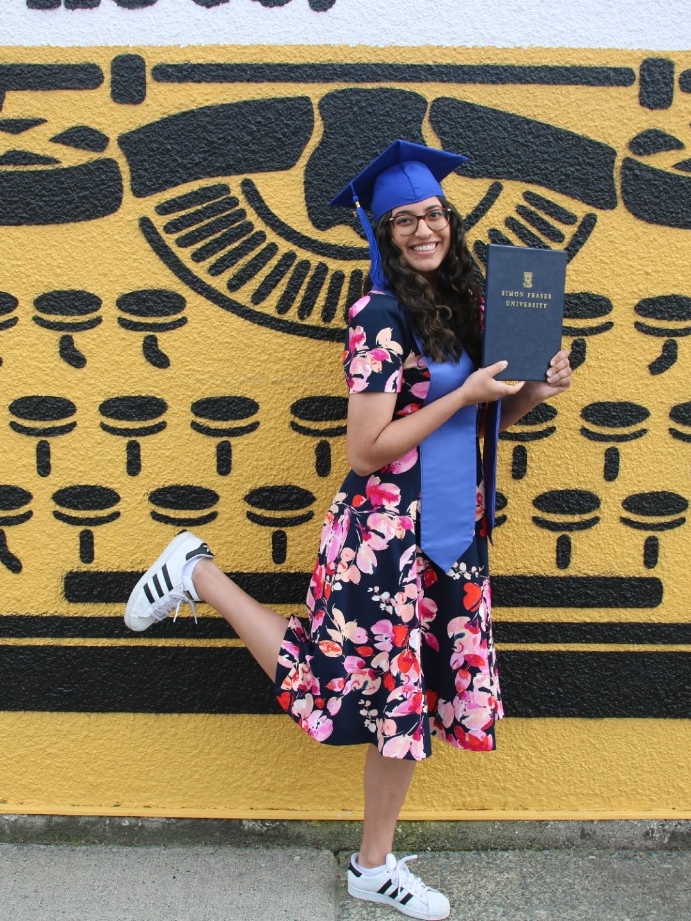 Nimrit Basra holding her diploma and wearing a graduation cap in front of a wall painted with a picture of a typewriter
