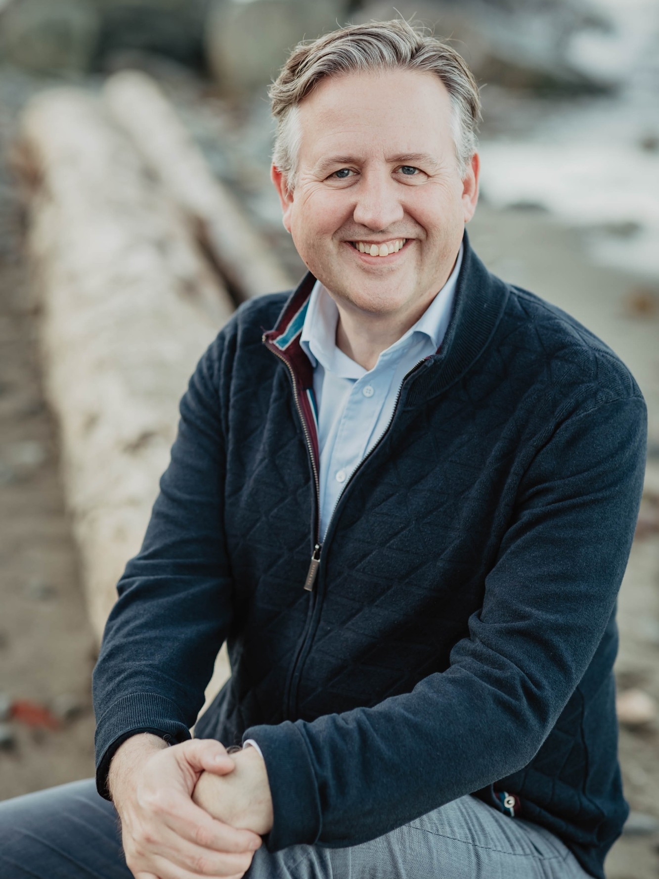 Kennedy Stewart sitting on a log on a beach, blurred in the background. He has gray hair and is smiling towards the camera. Kennedy is wearing a blue zip up coat with a blue button down underneath.