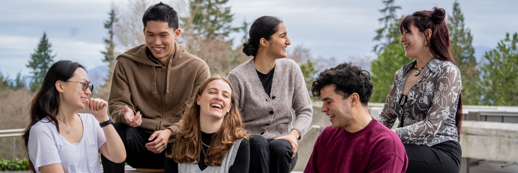 A group of six FASS Peer Mentors sitting on a bench interacting with each other. Some are smiling while others are laughing. They are photographed on SFU Burnaby Campus in the AQ with the mountains visible in the distance.