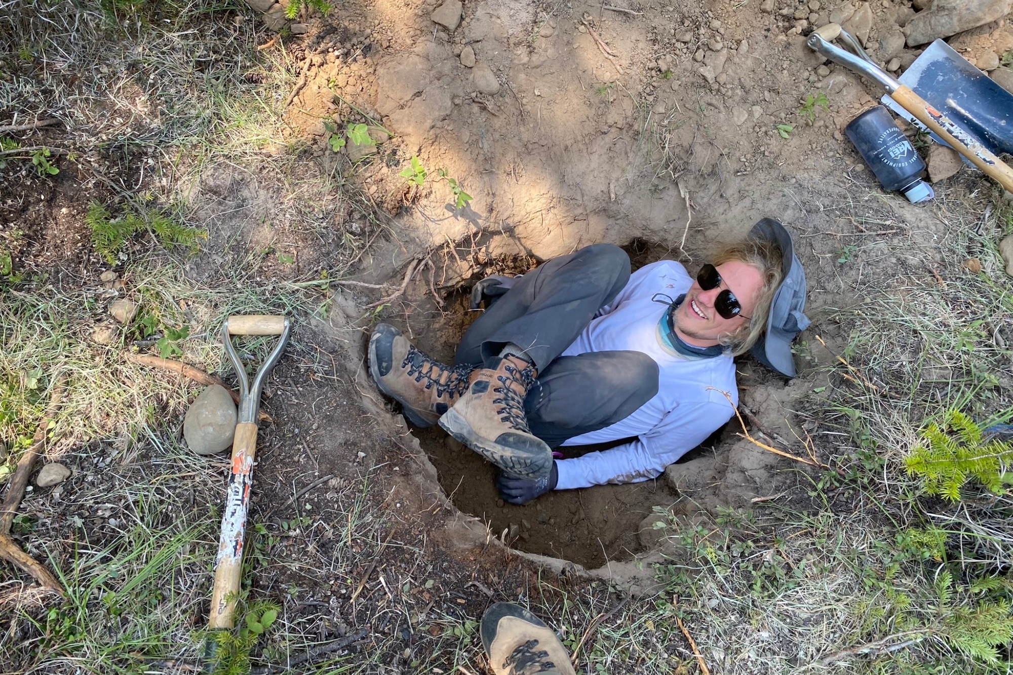 Matthew smiling in a hole in the ground next to a shovel