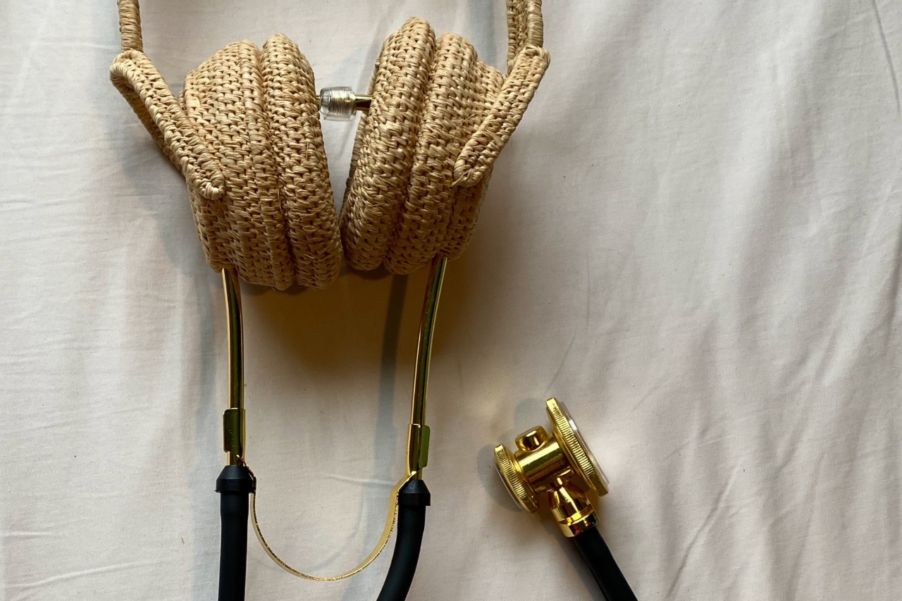 Against a simple white surface, a black and gold stethoscope. The ear tubes and ear tips are replaced by a palm fibre basket handwoven in the shape of life-sized over-the-ear noise cancelling headphones. This basket can serve to translate heartbeats into love songs. Light glints off of small gold cuffs along the rubber tubing of the stethoscope, and the tubing is gently curved so that the gold drum is set next to the woven headphones.