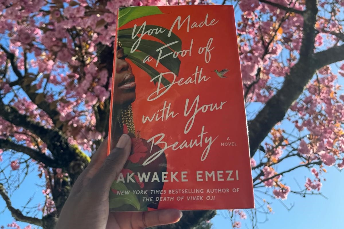 A hand holding up a copy of You Made a Fool of Death With Your Beauty by Akwaeke Emezi, with a cherry blossom tree in the background