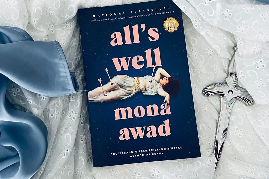The book, All’s Well by Mona Awad, lies on a bed, next to a sword, on top of blue satin and white lace fabric. The book cover is blue and depicts a woman shot with multiple arrows.  