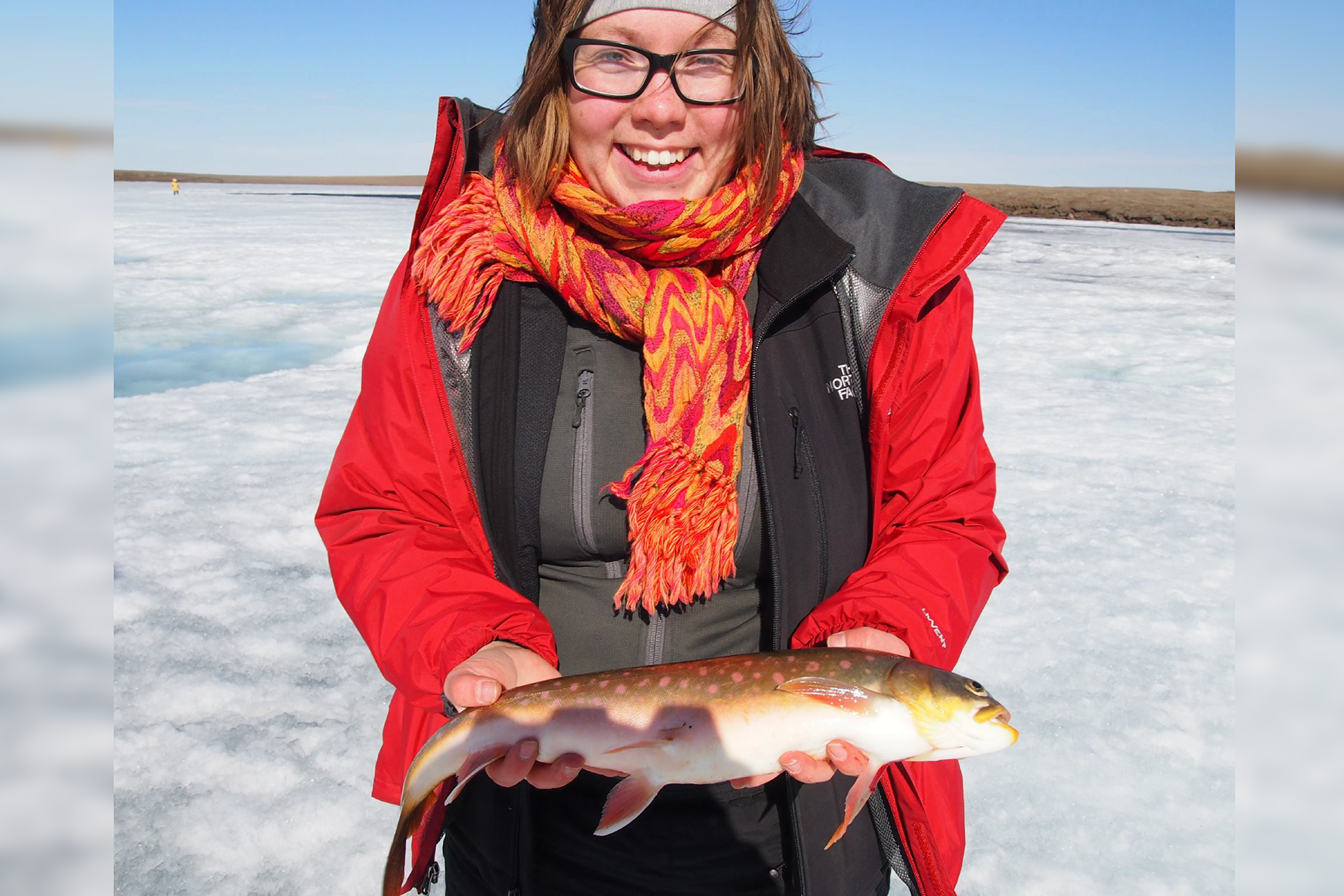 Zoe Todd standing in a winter landscape, wearing cold weather clothes, smiling and holding a fish