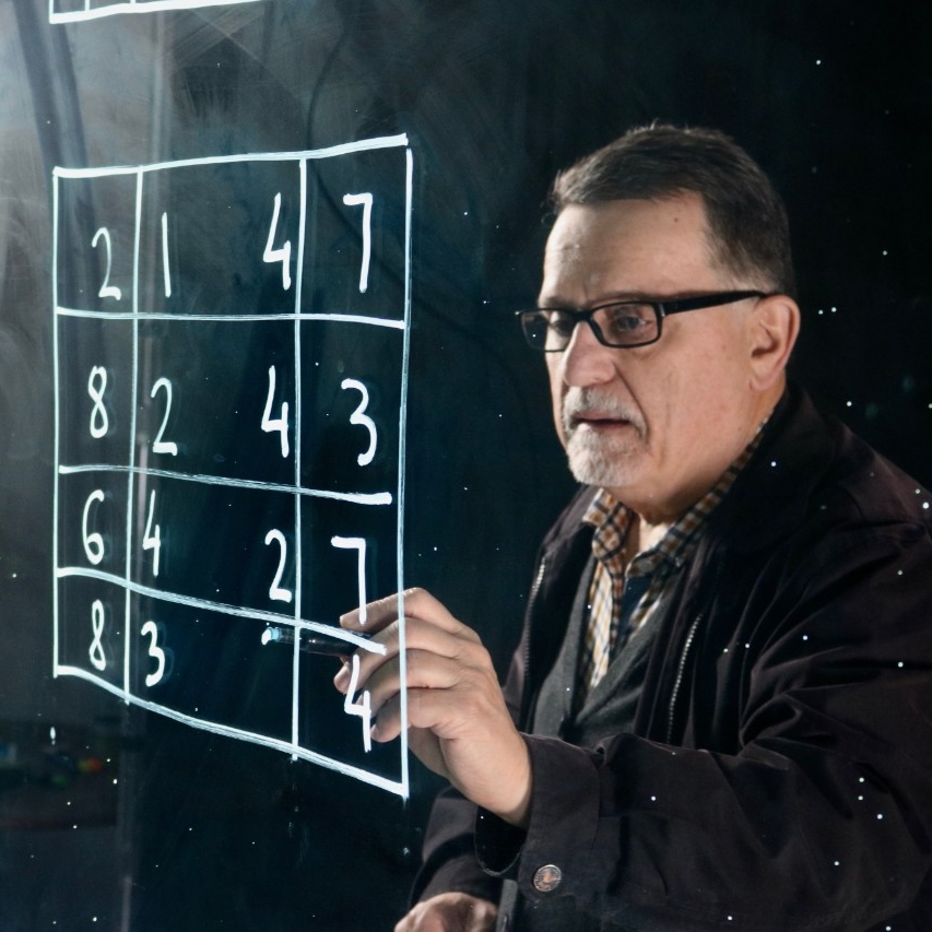 A photo of Veselin Jungic solving a puzzle with a writing tool. The puzzle is in white ink on a transparent surface. It consists of a square with intersecting lines and numbers between the lines. 