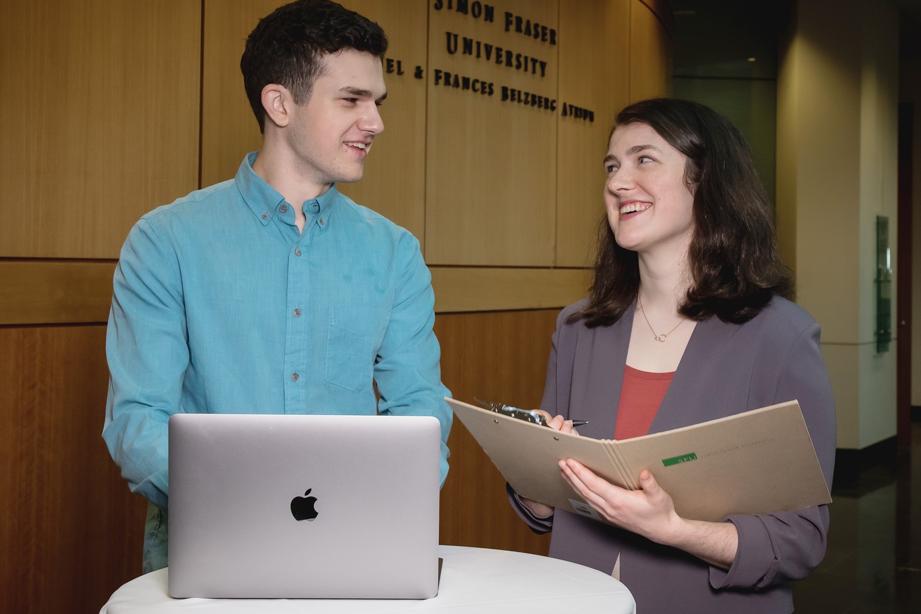 two undergraduate students smile at each other, one with an open laptop and another holding an open binder
