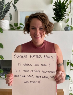 A woman holding a sign that reads: “Consent matters because it opens the door to a more positive relationship with yourself, your body and each other”