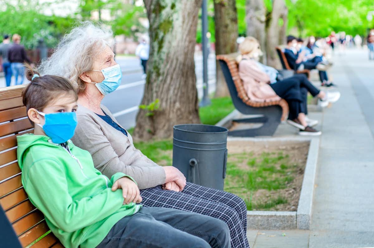 An older woman and a child sit on a park bench wearing masks.