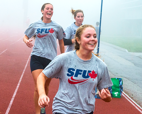 three girls in SFU shirts jogging on a track field