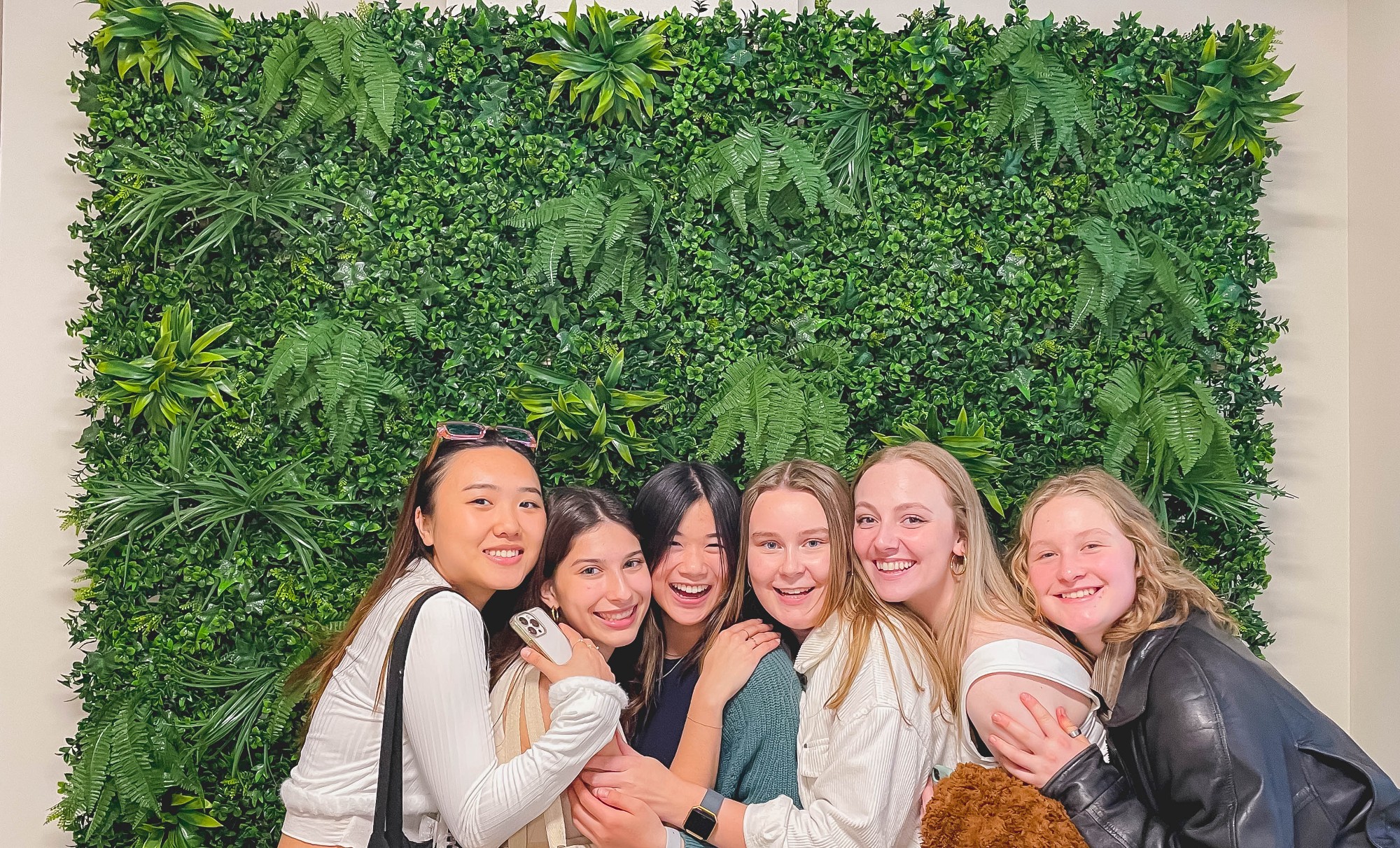 A group of female undergraduate students hanging out at a restaurant standing in front of a green leafy decor
