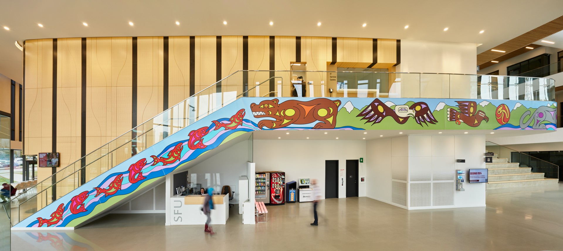 Indigenous artwork along a staircase in an open atrium. The artwork depicts a stream of fish meeting a bear. Next to it, there is are eagles soaring around the sun and a wolf. 