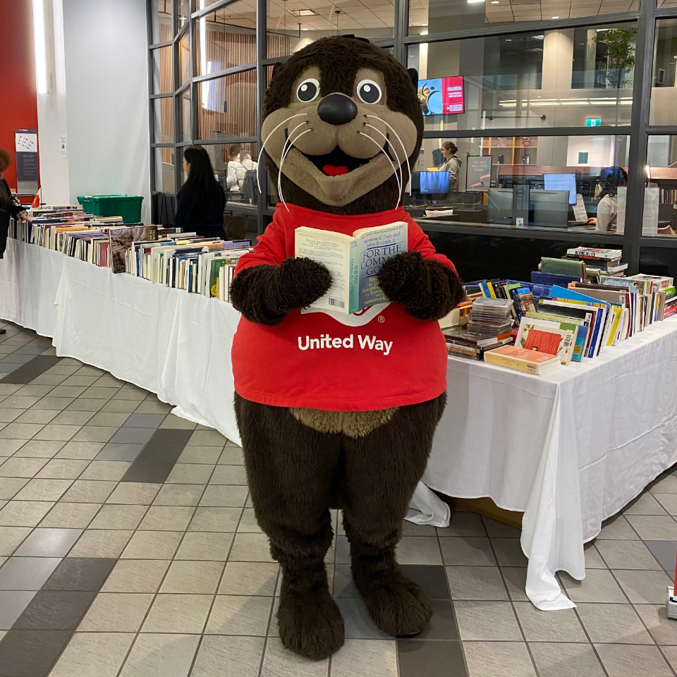United Way mascot, Seymour the Sea Otter, standing in front of a table full of books at the 2022 United Way Book and Bake Sale at Vancouver campus