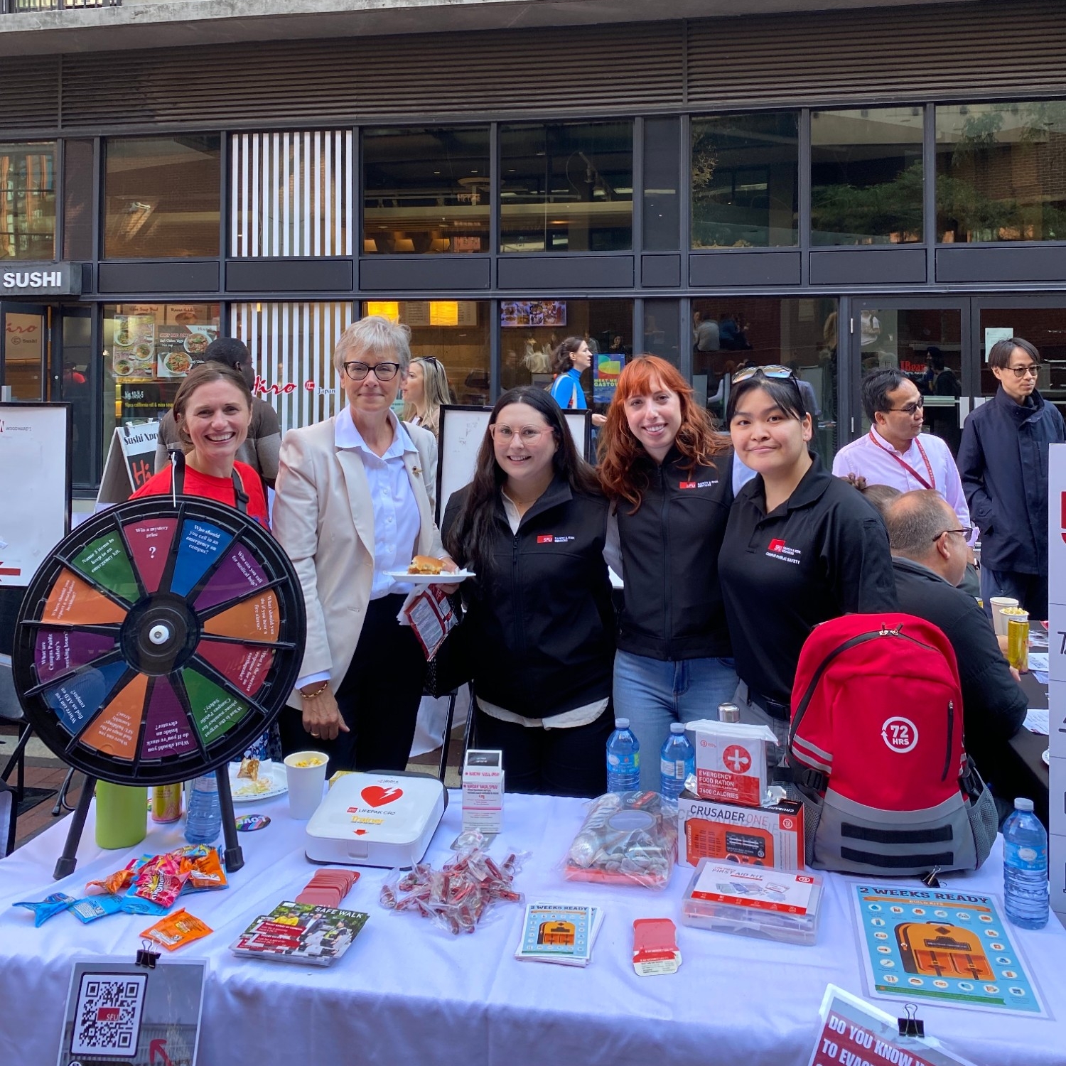 SFU President, Joy Johnson, with a group of SFU Safety & Risk Services staff standing behind a table at 2023 SFU Vancouver Welcome Back BBQ