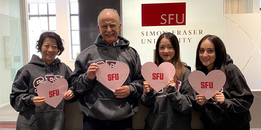 Four SFU Vancouver Admin team staff members wearing SFU hoodies and holding pink heart-shaped signs that say "SFU"