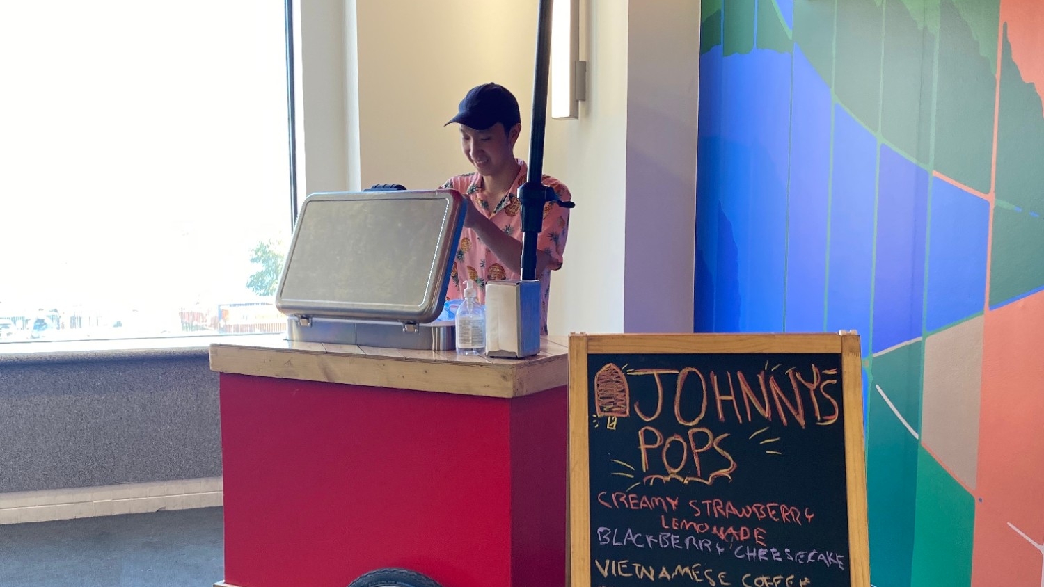 A Johnny's Pops staff member standing behind a cart with a sign that lists popsicle flavours at the 2023 Summer Social