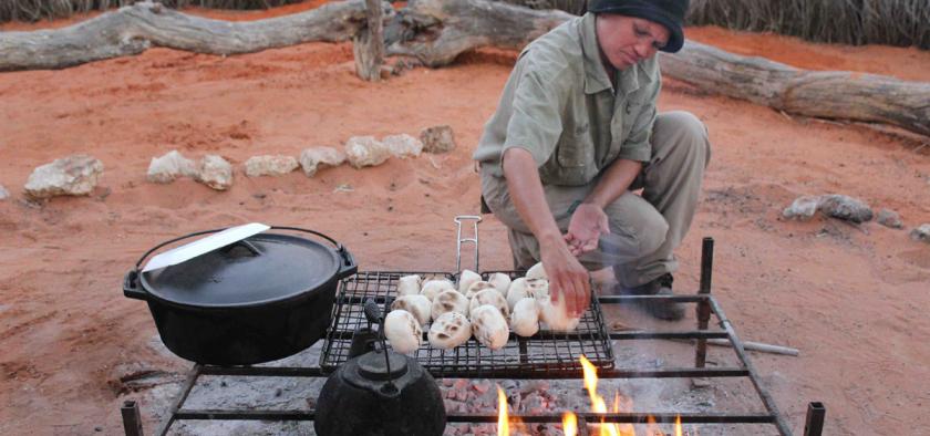 A +Khomani guide at //Uruke Bush Camp Adventures prepares bread buns on a grill 