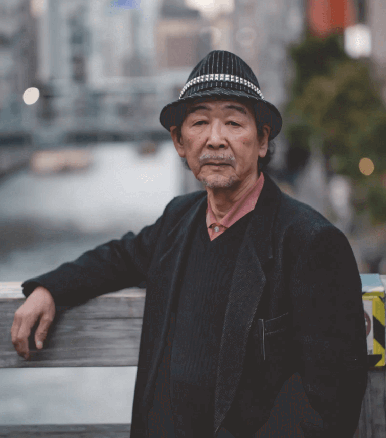 A portrait of a middle-aged Asian man wearing a black pin striped fedora, black blazer and sweater.