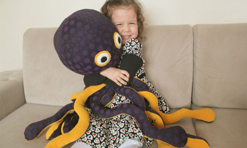 A happy little girl sitting on a beige sofa hugging a large purple spotted octopus plush with long dangly tentacles