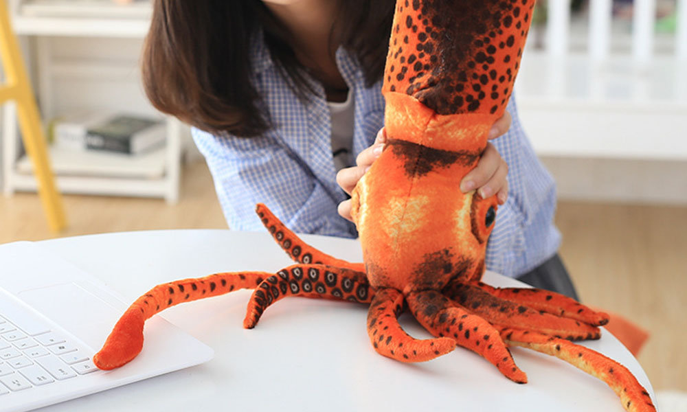 An orange with black spots squid plush held with two hands on a white table by a girl