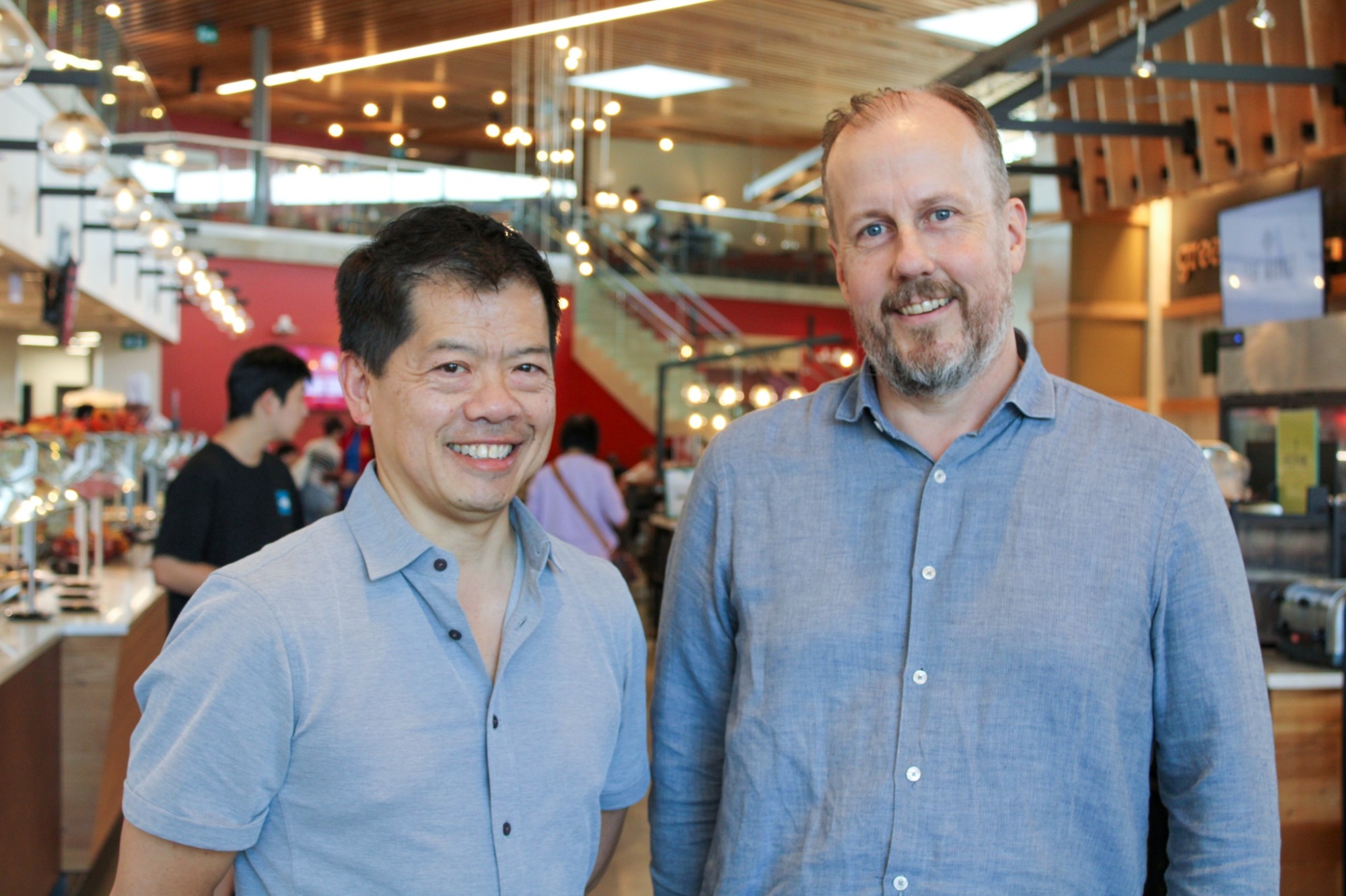Photo of two people standing side by side, one shorter wearing a blue short sleeved shirt, the other taller with a beard and blue long sleeved shirt. The interior Dining Commons at SFU can be seen in the background.