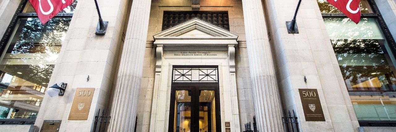 Front entrance of the SFU Beedie School of Business Segal Building in downtown Vancouver, featuring red SFU flags and stone columns. 