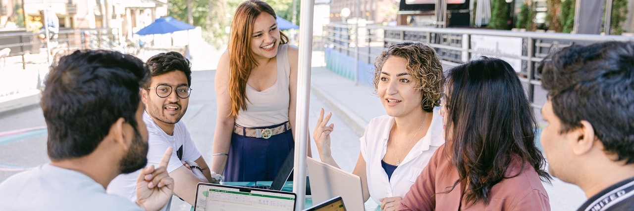 A group of young adults sitting outdoors around a table, talking and smiling while working on laptops.