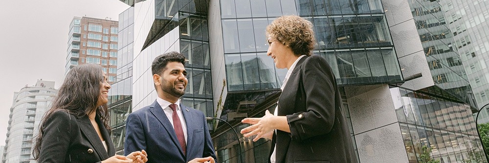 Two SFU Beedie graduate students and a faculty member are having a conversation outside a modern building.