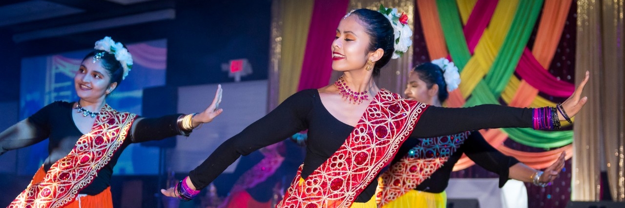 Three women dressed in black with red sashes and flowers in their hair dance at Diwali event.