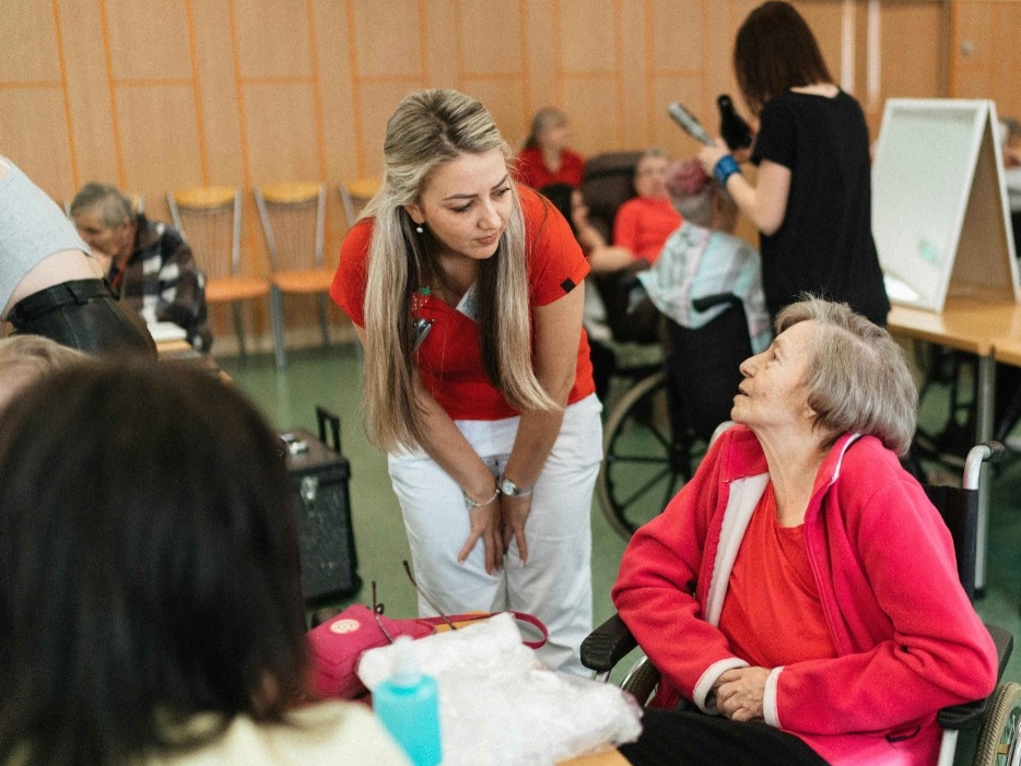 COPE Engage title written over a picture. The picture shows a workshop setting, an older woman in a orange sweater sits in a wheelchair, conversing with a younger woman. In the background, other older adults in wheelchairs, a working aisle, and another research assistant are visible.