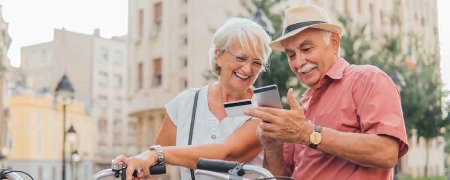 Smiling elderly couple using a smartphone while holding a credit card, standing outdoors with their bicycles in a city setting