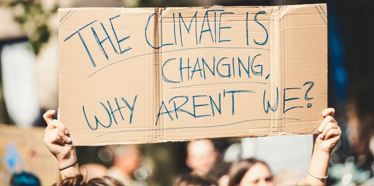 Cardboard banner with hand written statement saying 'Climate is changing, why aren't we' in a protest setting