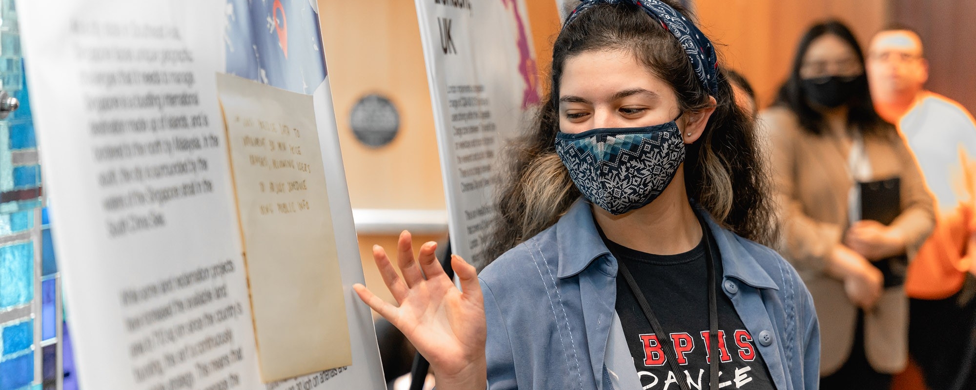 A person wearing a mask motions to their poster board at a conference