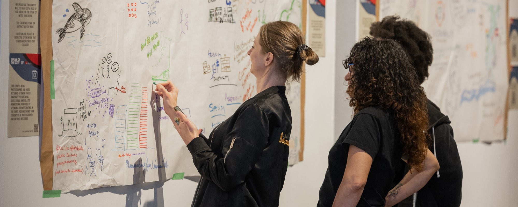 Two participants in a Hey Neighbour Collective dialogue watch another participant writing their thoughts on presentation paper on the wall. The paper is filled with thoughts and drawings from other participants. 