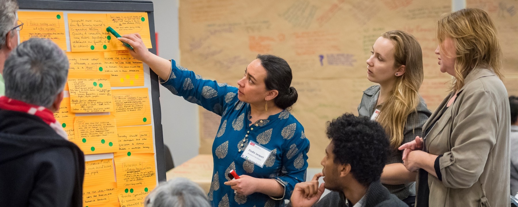 A facilitator points to a whiteboard covered in sticky notes as she speaks to dialogue participants