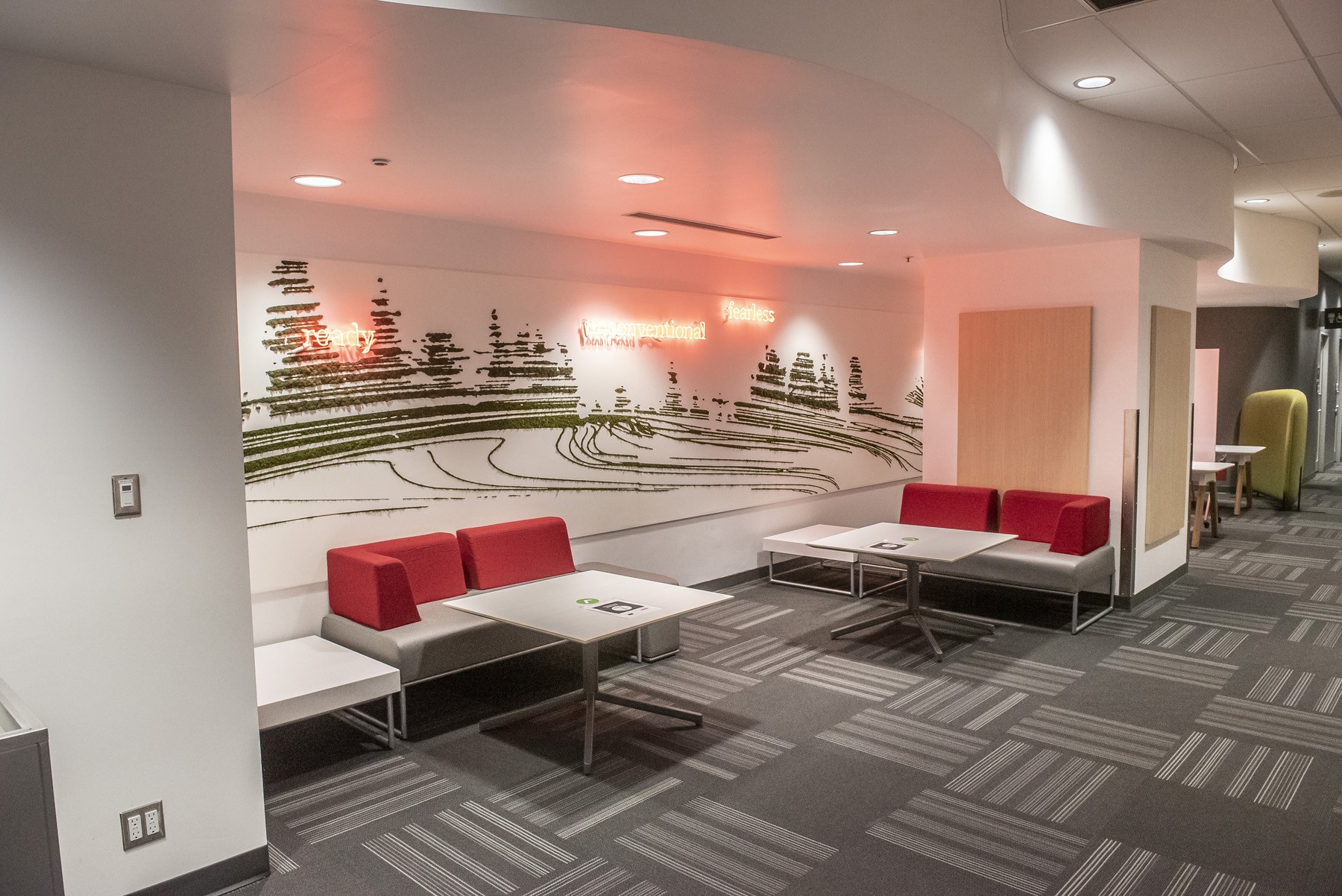 A study area at SFU's Harbour Centre campus. There are two red couches, each with a moveable table. A mural behind the couches depicts abstract black brush marks on a white background, resembling trees, with the words "ready", "unconventional" and "fearless" in red neon lights.