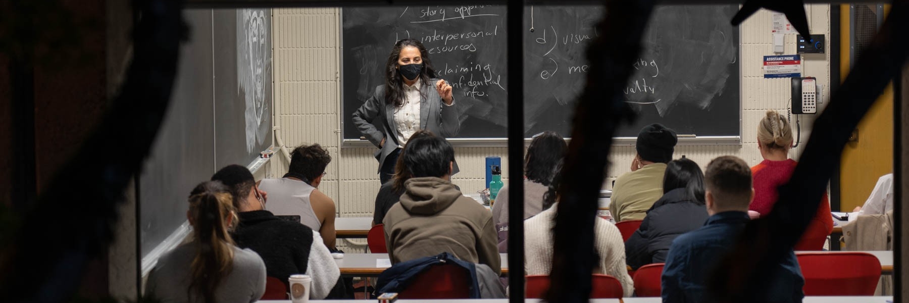 teacher stands in front of a blackboard lecturing to a room full of students