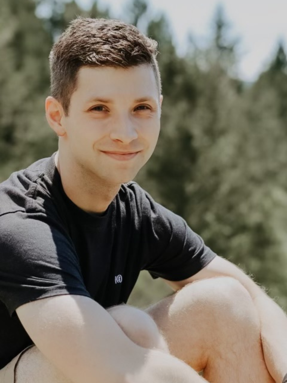 Jason is sitting outside wearing a black t-shirt smiling at the camera. In the background are green plants that are slightly blurred.