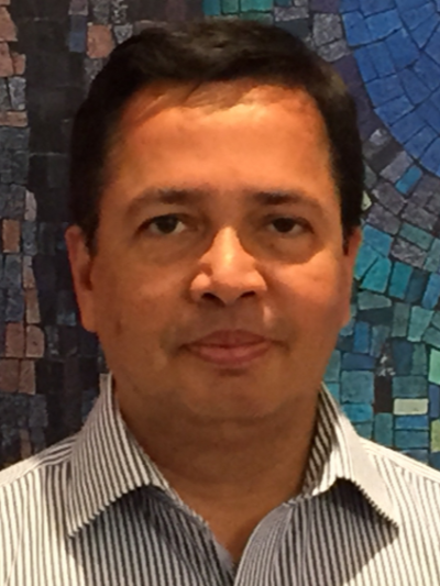 Dr. Habib Chaudhury is photographed in front of the blue SFU mosaic on the Burnaby campus. He is wearing a striped collared shirt and has short dark hair.